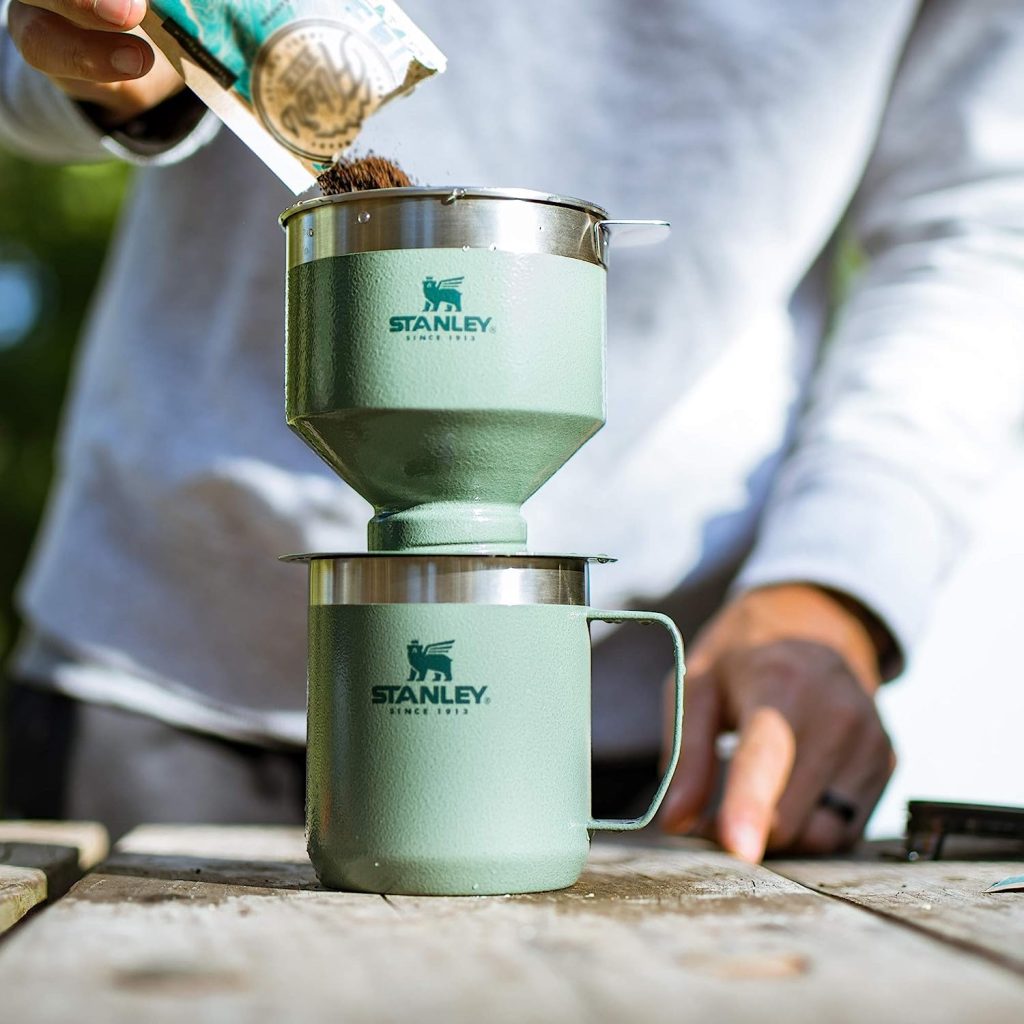 Adventurer using the Stanley Pour Over Set to brew coffee by a campfire, showcasing its portability and outdoor utility.