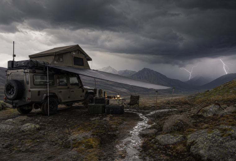 Storm-ready overland campsite on high ground with secured shelter, waterproof gear, and dark storm clouds approaching.
